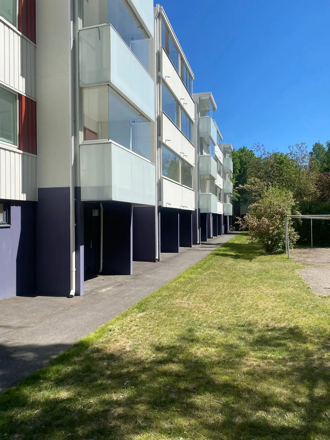 Pitkä, moderni kerrostalo vaalealla julkisivulla ja tummansinisellä sokkelilla. Rakennuksessa on useita parvekkeita lasikaiteilla, joissa on osittain himmeää lasia. Vasemmassa päädyssä on punaisia pystypaneeleja. Rakennuksen vieressä kulkee asfaltoitu kävelytie ja vihreä nurmikko, jolla näkyy varjoja. Taustalla on puita ja pensaita kirkkaan sinisen taivaan alla.
