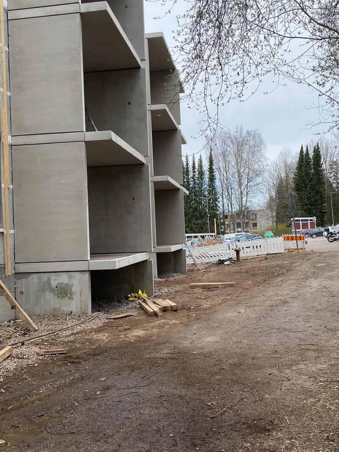 Harmaa betonirakennus rakenteilla, jossa on useita ulkonevia kerroksia tai parvekkeita. Etualalla on mutainen maapolku, jossa on soraa, puulautoja ja rakennusjätettä. Taustalla näkyy rakennustyömaa valkoisine aitoineen, autoja, moottoripyörä ja puita. Taivas on pilvinen ja valaistus tasainen.