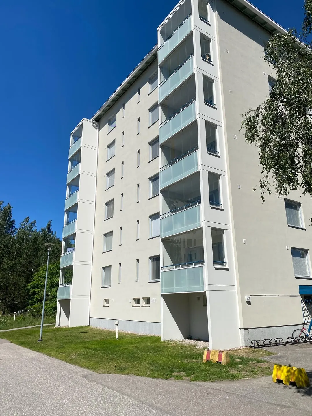 Vaaleanharmaa monikerroksinen asuinrakennus, jossa on lasitetut parvekkeet, seisoo kirkkaan sinisen taivaan alla aurinkoisena päivänä. Rakennuksen edessä on vihreä nurmikko ja asfalttitie. Vasemmalla näkyy puita ja katuvalo, oikealla puun lehvästöä. Alhaalla oikealla on polkupyörätelineitä, punainen polkupyörä ja keltaisia sekä punaisia betoniporsaita.