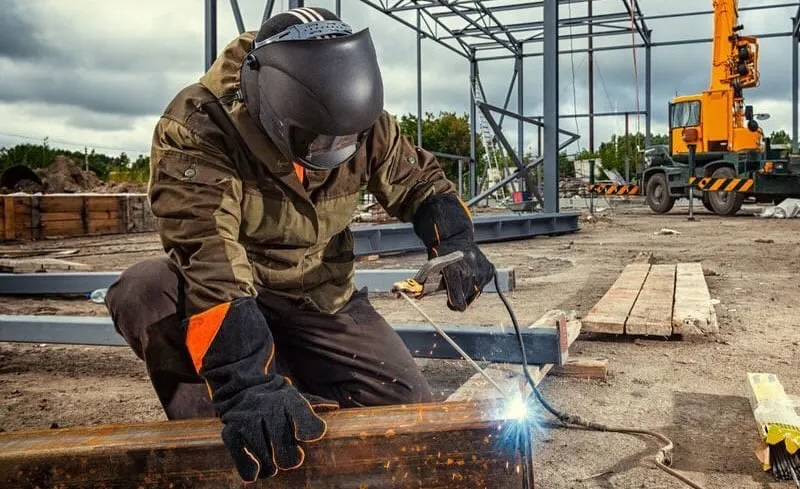 Hitsaaja polvillaan rakennustyömaalla hitsaamassa suurta metallipalkkia. Hänellä on yllään ruskea takki, tummat housut, musta hitsauskypärä ja musta-oranssit hitsauskäsineet. Hitsauskohdasta nousee kirkkaita sinisiä kipinöitä ja valoa. Taustalla näkyy harmaa metallirunko, keltainen liikkuva nosturi ja pilvinen taivas.