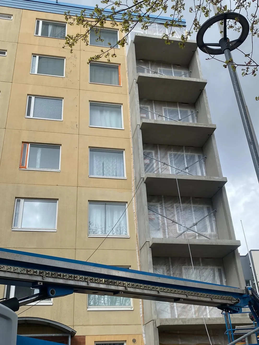 Korkea rakennus, jossa on keltainen julkisivu vasemmalla ja harmaa betonirakenne oikealla. Betonirakenteen tasot on peitetty läpinäkyvällä muovilla ja ristiin asetetuilla metallitangoilla. Kuvan alareunassa on sinisen rakennusnosturin varsi valoineen. Yläosassa näkyy puun oksa vihreine lehtineen ja oikealla puolella katulamppu pyöreällä yläosalla. Taivas on pilvinen ja valaistus hajavaloa.