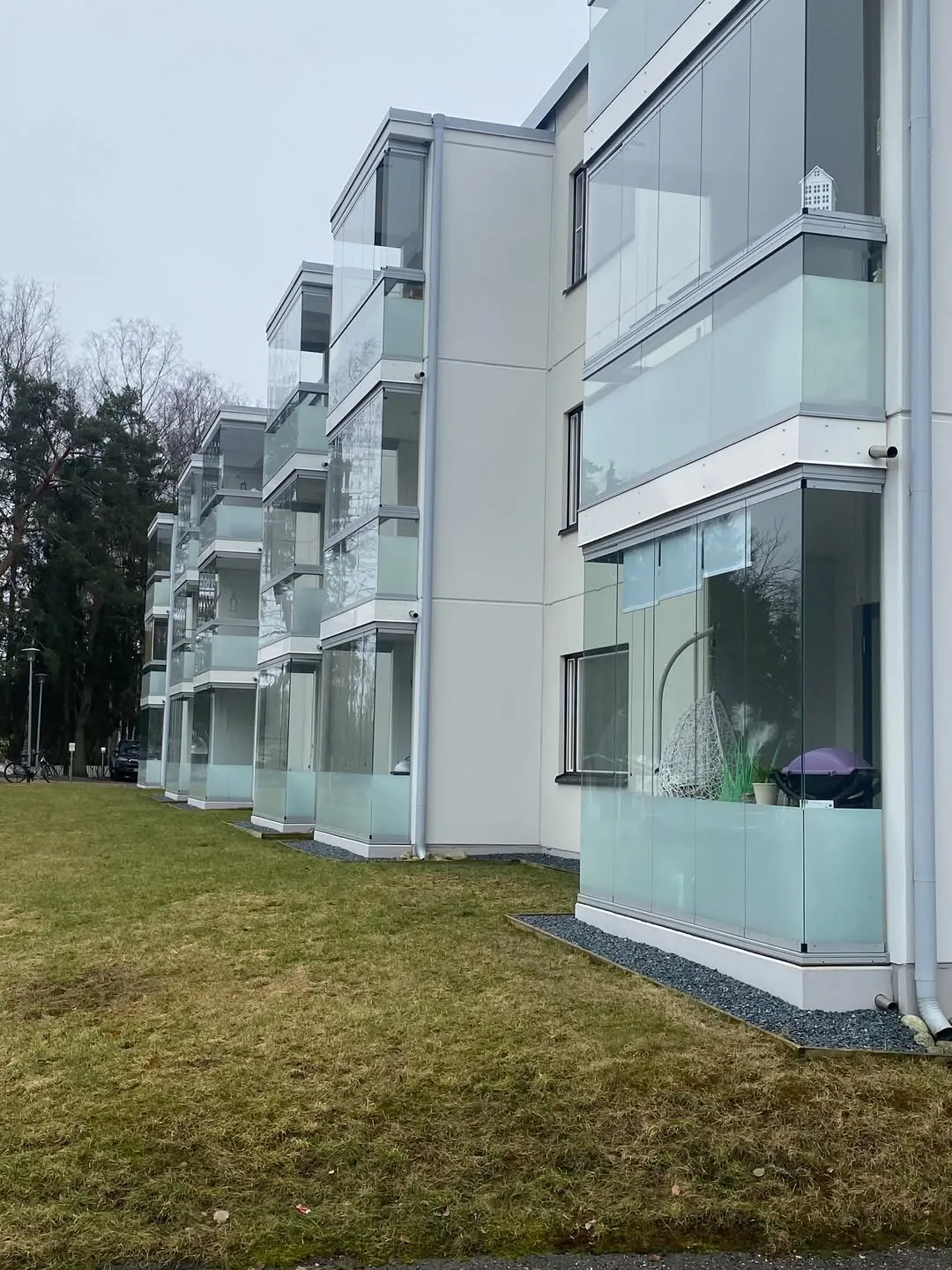 Rivi moderneja, vaaleita kerrostaloja lasitetuilla parvekkeilla. Rakennusten edessä on tummanharmaa sorakaistale ja laikukas vihreä nurmikko. Taustalla näkyy puita ja pilvinen harmaa taivas. Yhdellä parvekkeella on valkoinen riipputuoli ja violetti grilli.