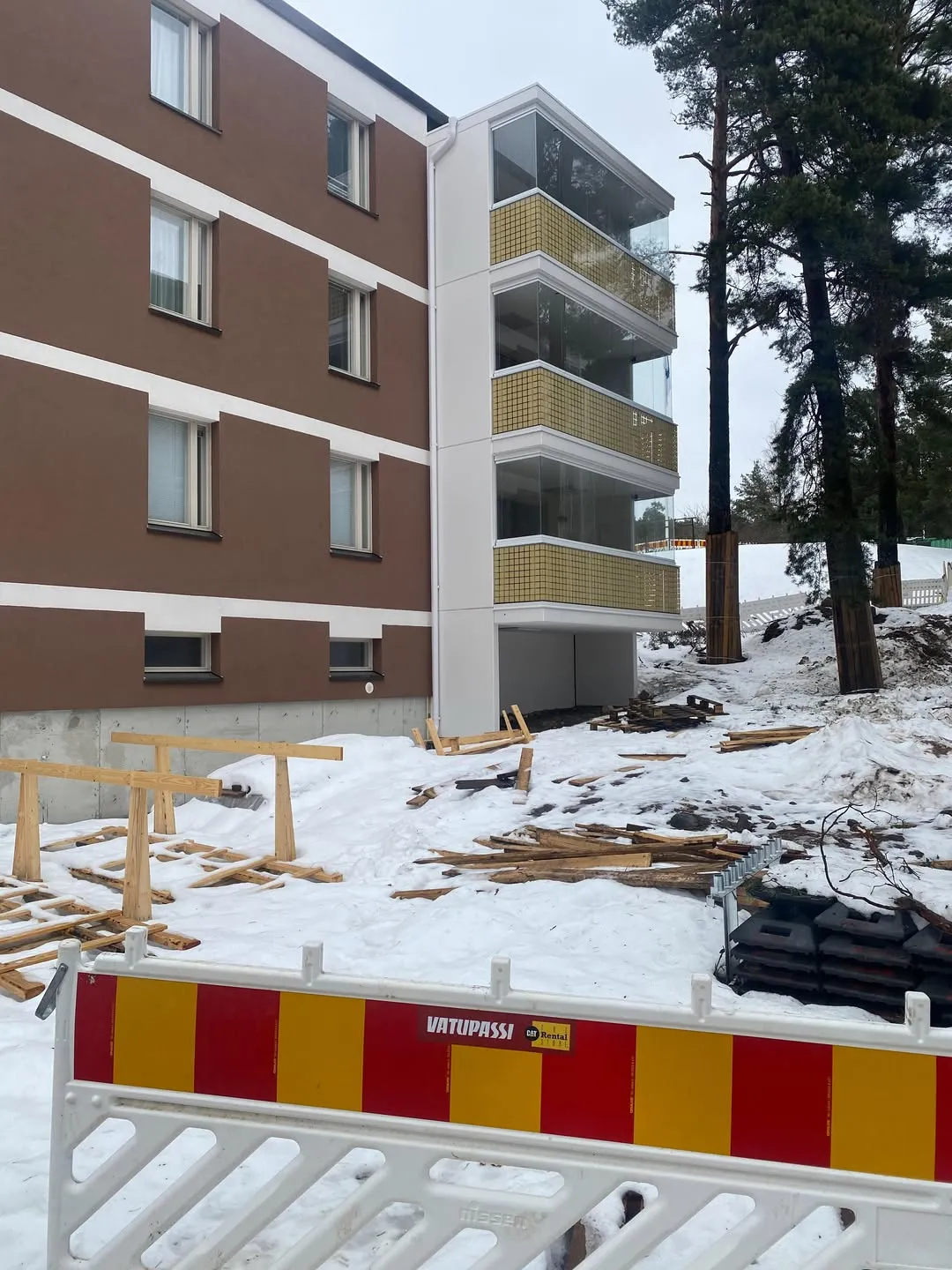 Ruskea kerrostalo valkoisilla vaakaraidoilla ja ikkunoilla. Rakennuksen oikealla puolella on kolme lasitettua parveketta keltaisilla kaiteilla. Etualalla on valkoinen, punakeltainen rakennusaita, jossa lukee 'VATUPASSI' ja 'CAT Rental'. Maassa on lunta ja puutavaraa, ja taustalla näkyy mäntyjä pilvisenä päivänä.
