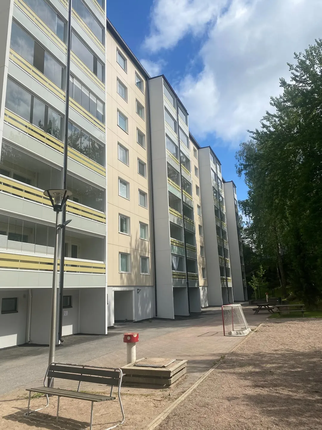Rivi vaaleankeltaisia kerrostaloja, joissa on lasitetut parvekkeet keltaisilla kaiteilla, seisoo aurinkoisena päivänä. Rakennusten edustalla on kivetty ja hiekkainen piha-alue, jossa on penkki, punainen sieni-esine, pieni jalkapallomaali ja piknikpöytiä. Oikealla puolella kasvaa tiheästi vihreitä puita sinistä, pilvistä taivasta vasten.