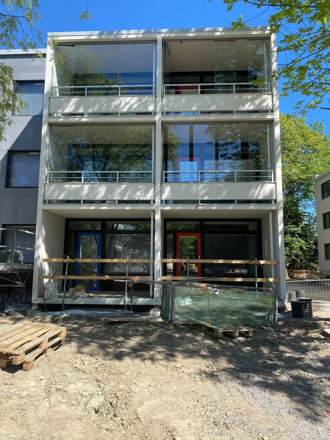 Aurinkoisena päivänä otettu kuva modernista, monikerroksisesta rakennuksesta. Rakennuksen valkoisessa julkisivussa on kaksi ylintä kerrosta, joissa on suuret lasitetut parvekkeet, jotka heijastavat puita ja sinistä taivasta. Maatasolla on rakennustyömaa, jossa on kaksi sisäänkäyntiä: toisessa on sininen ja toisessa punainen ovenkarmi. Edustalla on puulavoja ja pino lasilevyjä. Maassa näkyy varjoja puista.