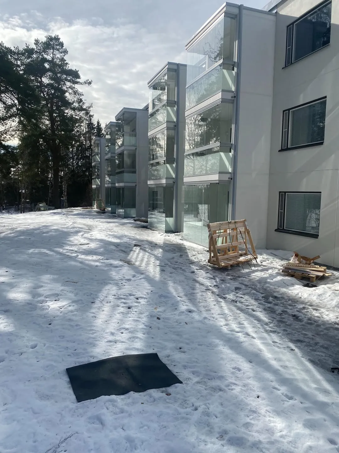 Moderni kerrostalo, jossa on lasitetut parvekkeet, seisoo lumisessa maisemassa. Maassa on lunta, jalanjälkiä, tumma matto ja puulavoja. Taustalla näkyy tiheä metsä. Aurinkoinen sää luo pitkiä varjoja lumeen.