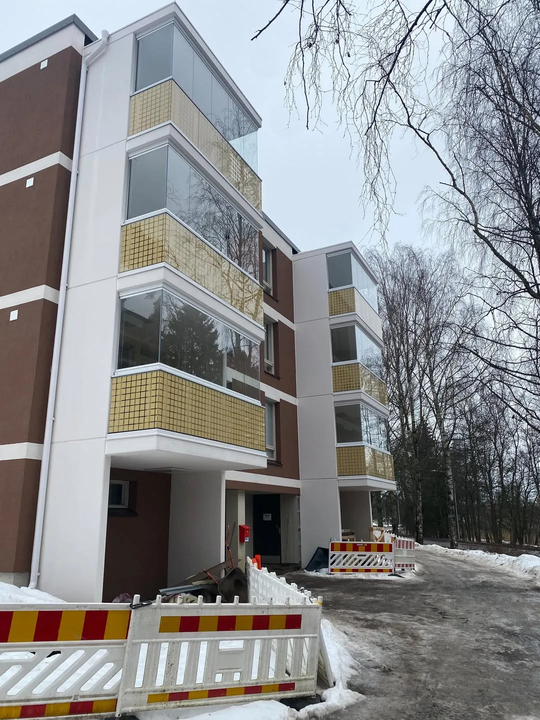 Monikerroksinen asuinrakennus, jossa on ruskeat seinät ja valkoiset pilarit. Lasitetuissa parvekkeissa on kultaiset ruudukkokuvioiset kaiteet. Edustalla on valkoisia, puna-kelta-raidallisia rakennusaitoja ja lunta. Taustalla on paljaita puita pilvisenä talvipäivänä.