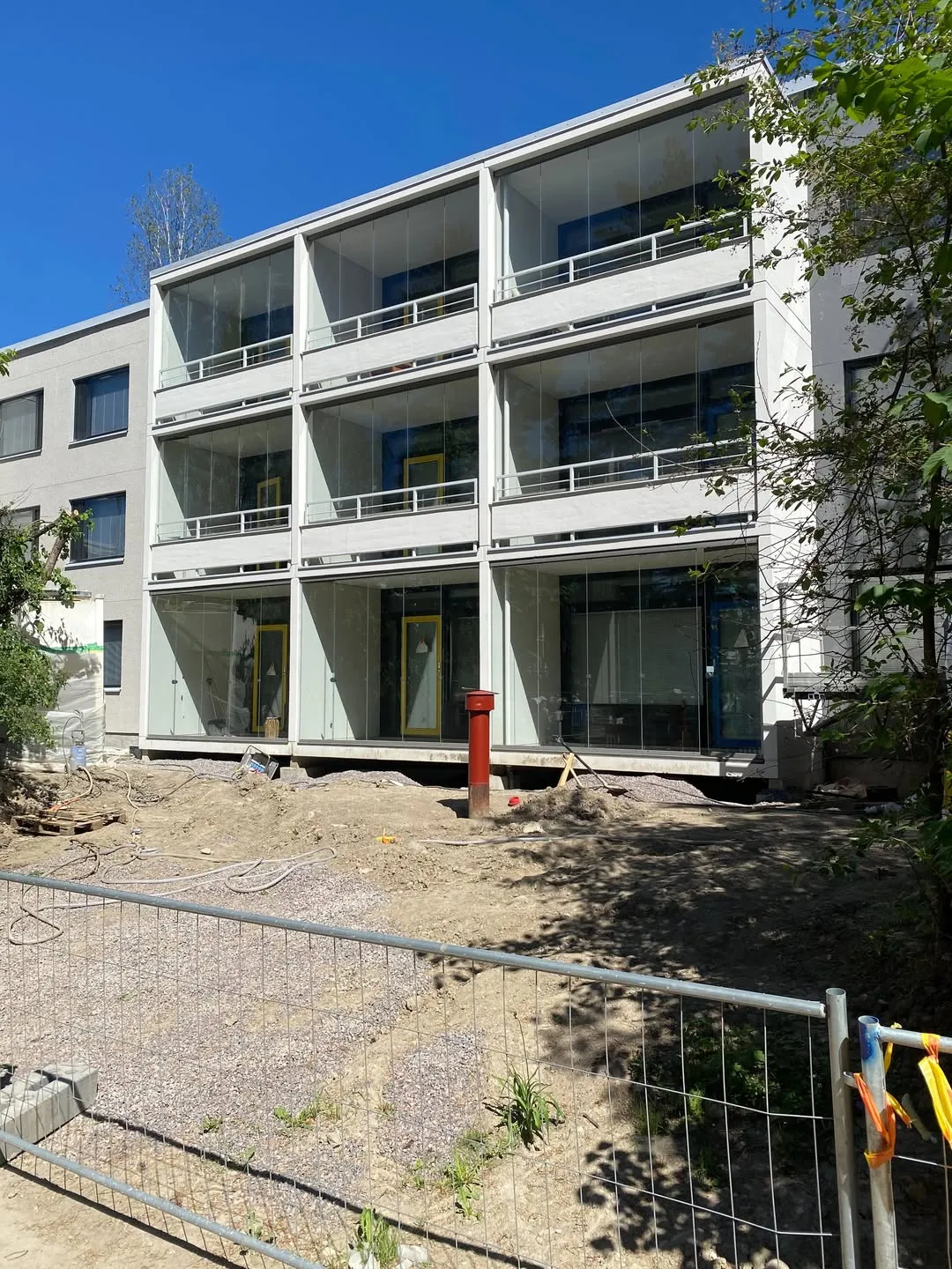 Aurinkoisena päivänä otettu kuva vaaleasta asuinrakennuksesta, jossa on kolme kerrosta lasitettuja parvekkeita valkoisine kaiteineen. Parvekkeiden sisällä näkyy keltaisia ikkunan- tai ovenkarmeja. Rakennuksen edessä on rakennustyömaa-alue, jossa on paljasta maata, soraa ja punainen pystysuora putki. Etualalla on metalliverkkoaita. Taustalla on kirkas sininen taivas ja vihreitä puita.