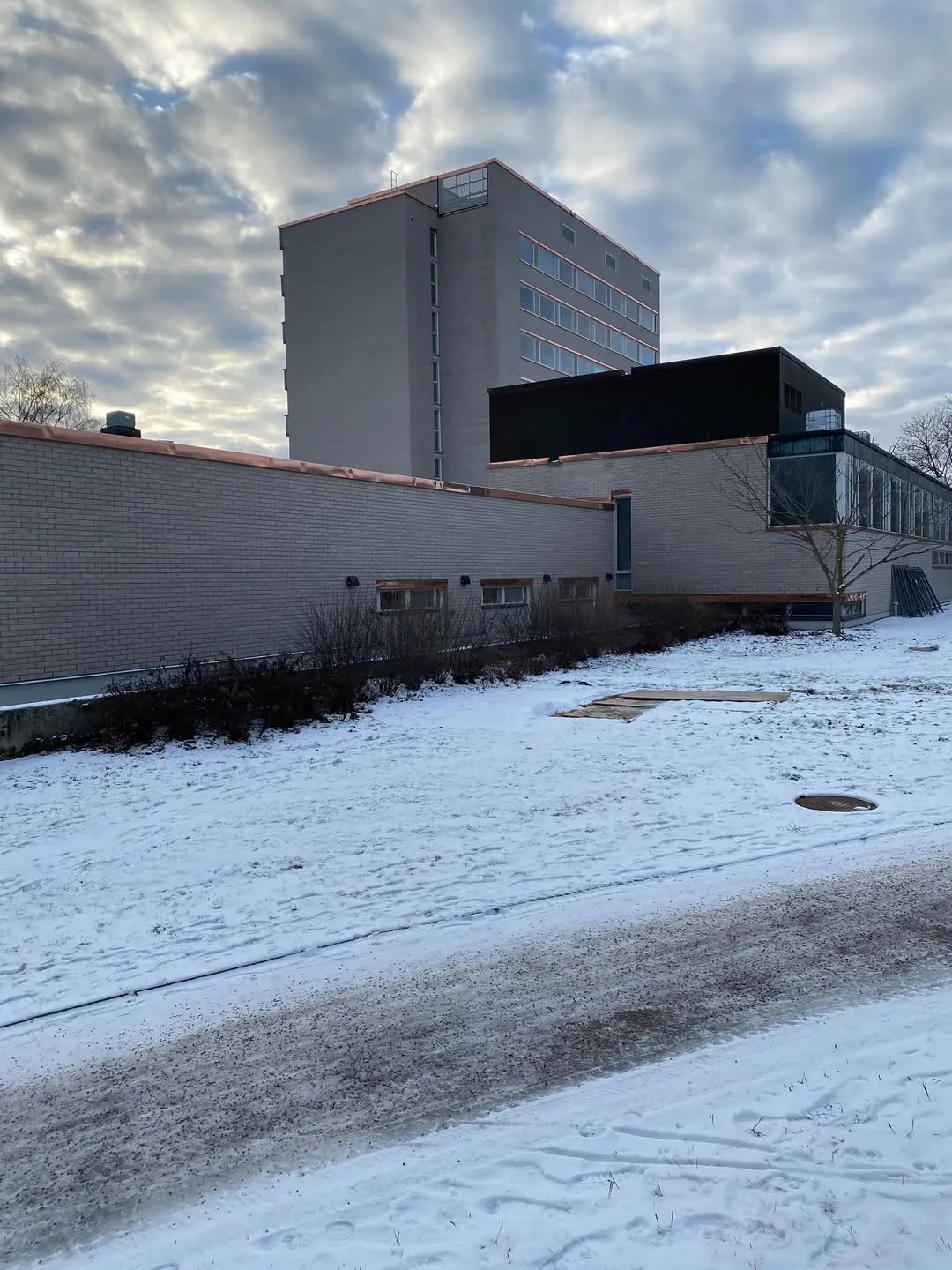 Talvinen näkymä, jossa etualalla on lumipeitteinen maa ja osittain sulanut tie. Taustalla kohoaa korkea, harmaa rakennus, jossa on useita ikkunoita ja kuparinvärisiä yksityiskohtia. Sen edessä on matalampi, harmaatiilinen rakennus, jonka katolla on kuparinväriset reunalistat. Oikealla puolella matalammassa rakennuksessa on suuria lasipintoja ja musta yläosa. Taivas on pilvinen.